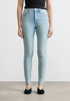 Femme portant un jean skinny taille haute bleu clair associé à des ballerines noires et un haut sans manches noir, debout sur un sol gris.