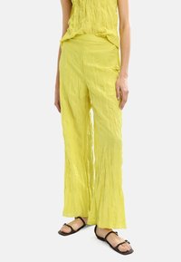 Jasnożółte, marszczone spodnie o szerokich nogawkach z teksturowanym wykończeniem. Posiadają płaski waistband i luźny krój, zestawione z czarnymi sandałami na paski.