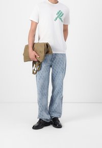 T-shirt blanc avec texte vert, jean bleu clair à motifs et chaussures noires. Un sac beige est tenu dans la main gauche.