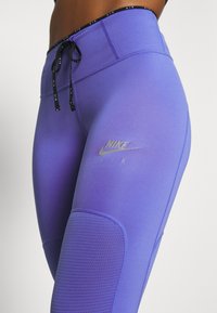 Nike legíny v gradientní fialové barvě, s vysokým pasem, černou šňůrkou na stažení a síťovanými panely pro větrání. Součástí je stříbrné logo Nike Air.