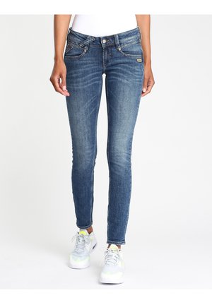 Personne portant un jean bleu slim fit avec des détails délavés et des baskets blanches, debout devant un fond clair uni.
