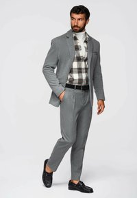 Grå blazer med subtil textur, kombinerad med en rutad skjorta i vitt, svart och brunt, samt grå byxor. Svarta skor fulländar outfiten.