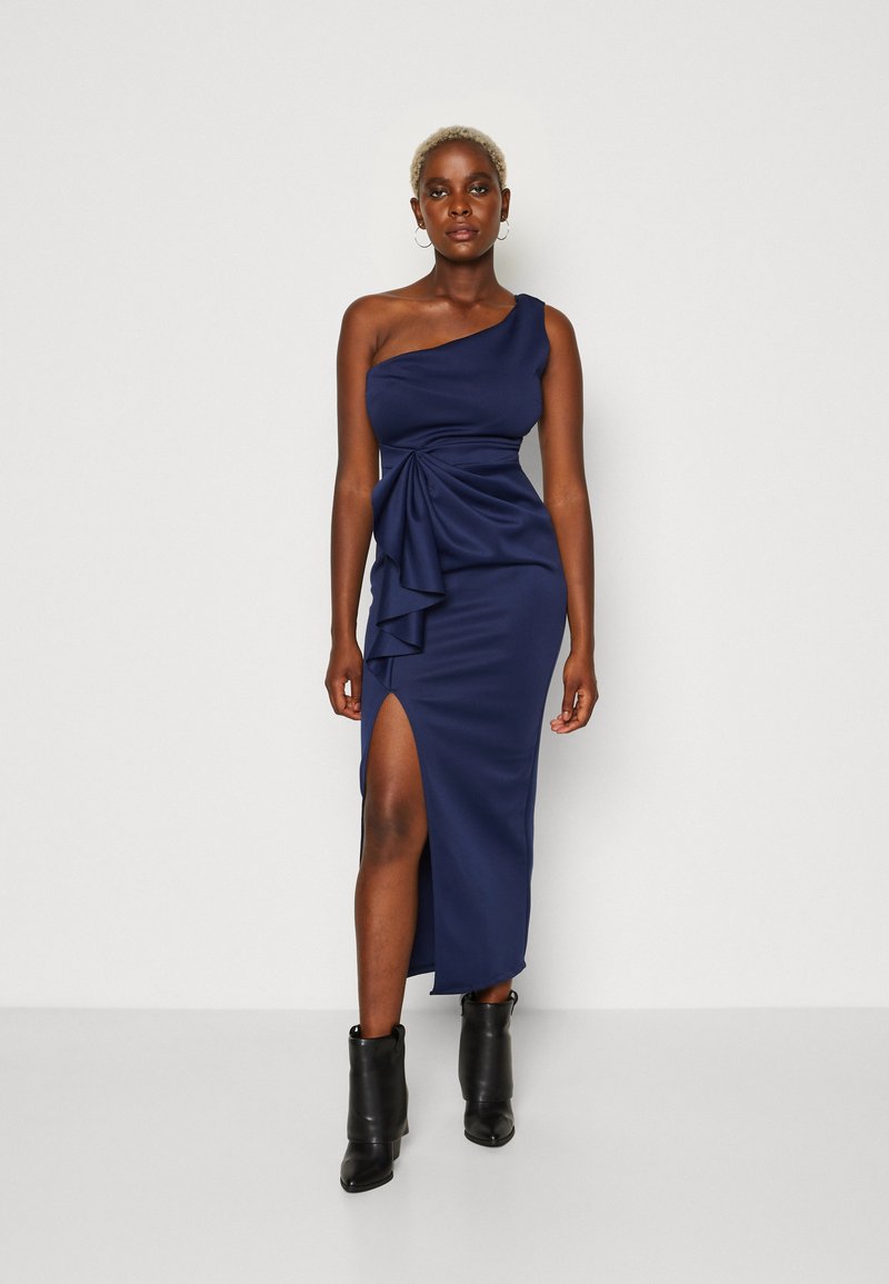 Robe bleu marine à une épaule en tissu lisse, avec un devant drapé, une fente latérale et un détail froncé à la taille. Assortie avec des bottines noires.