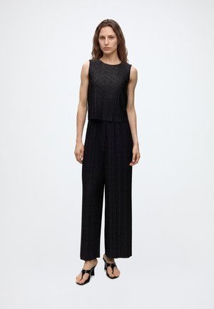 Femme portant un haut noir texturé sans manches et un pantalon large assorti, avec des sandales à talons noires ouvertes, debout devant un fond uni.