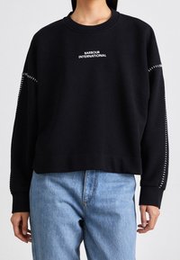 Černá mikina s dlouhými rukávy, vroubkovanými manžetami a kulatým výstřihem. Obsahuje bílé logo "Barbour International" a kontrastní bílé švy na rukávech.