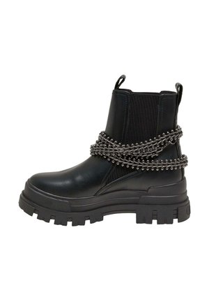 Jambes croisées portant des bottines noires épaisses avec un détail chaîne et une jupe noire brillante, assise sur un trottoir en milieu urbain.