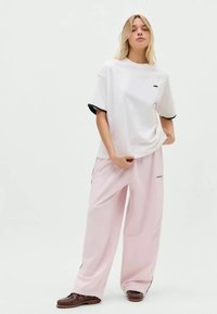 Una donna bionda indossa una t-shirt bianca oversize, pantaloni palazzo rosa chiaro e mocassini marroni su uno sfondo chiaro e semplice.