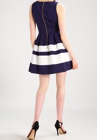 Mekko ilman hihoja, tummansiniharmaa, jossa on levenevä hameosa, vaakasuoria valkoisia raitoja ja näkyvä takavetoketju. Mustat korkokengät.