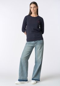 Maglione lavorato a maglia blu notte con collo rotondo e maniche lunghe, abbinato a jeans a gamba larga blu chiaro e sneakers bianche. Dettaglio accentato in rosso presente.