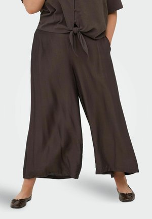 Žena nosí voľné tmavohnedé nohavice širokých nohavíc spojené so zodpovedajúcou predne zaväzovanou košeľou a tmavohnedými balerínami.