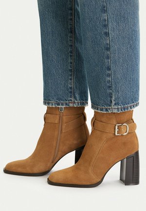 Braune Wildleder-Stiefeletten mit seitlichem Reißverschluss, dekorativem Riemen und silberner Schnalle, getragen zu blauen Jeans.