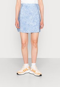 Henkilö, joka on pukeutunut sinivalkoiseen marmoroituun mini-hameeseen, valkoisiin sukkiin ja beigeihin lenkkareihin, joissa on oranssit raidat, seisoo harmaalla lattialla.