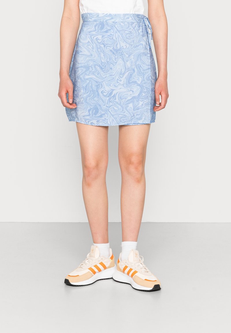 Henkilö, joka on pukeutunut sinivalkoiseen marmoroituun mini-hameeseen, valkoisiin sukkiin ja beigeihin lenkkareihin, joissa on oranssit raidat, seisoo harmaalla lattialla.