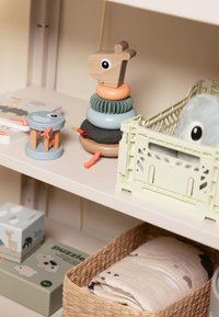 Jouet empilable en bois avec une tête de girafe, des anneaux multicolores (rose, bleu, vert) et une base texturée. Autres jouets et un panier tissé à proximité.