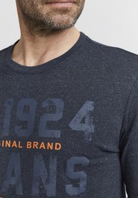 Tamsiai mėlyna marškinėliai iš tekstūruoto audinio su taškuoto rašto, dideliais spausdintais skaičiais "1924" ir "ORIGINAL BRAND" oranžine spalva.