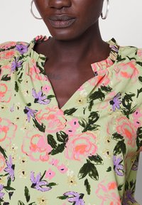 Blouse fleurie avec une base verte ornée de fleurs roses et violettes, un col en V et un détail froncé au niveau de l'encolure.