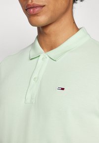 Lysegrønn polo skjorte med teksturert stoff, med en to-knapp placket og brodert logo i rødt, hvitt og blått på venstre bryst.