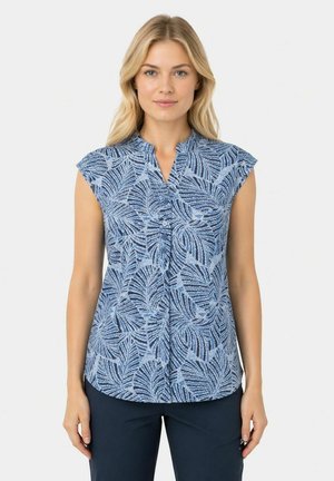 Frau trägt eine ärmellose blaue Bluse mit hellblauem Blattmuster und marineblaue Hose, steht vor einem einfarbigen hellen Hintergrund.