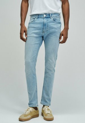 Hombre con vaqueros ajustados de color azul claro y zapatillas beige, de pie contra un fondo liso de color gris claro.