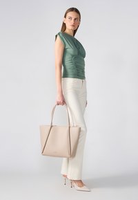 Beige läder toteväska med dubbla handtag, slät yta och guldlogotyp; kombineras med en grön topp och ljusa slim-fit byxor.