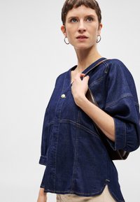 Σκούρο μπλε τζιν πουκάμισο με V-λαιμό, μεγάλο κουμπί και μανίκια τριών τετάρτων. Σημαντική αντίθεση ραφής και άνετη εφαρμογή.