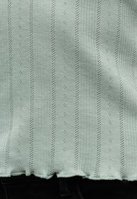 Ανοιχτό πράσινο πλεκτό ύφασμα με κάθετα ριμπ και σχέδια με οπές, κυματιστό τελείωμα, φορεμένο πάνω από μαύρο παντελόνι.