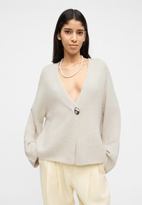 Νέα γυναίκα που φοράει ανοιχτό μπεζ ζακέτα με κουμπιά, κολιέ με πέρλες και κρεμ πτυχωτό παντελόνι, στέκεται με τα χέρια στις τσέπες.