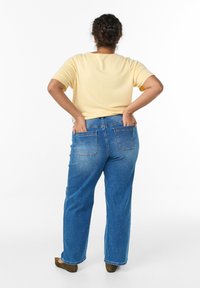 Žltý top s krátkymi rukávmi, modré rifle s vysokým pásom a zadnými vreckami, priamy strih, a hnedé topánky s okrúhlou špičkou.