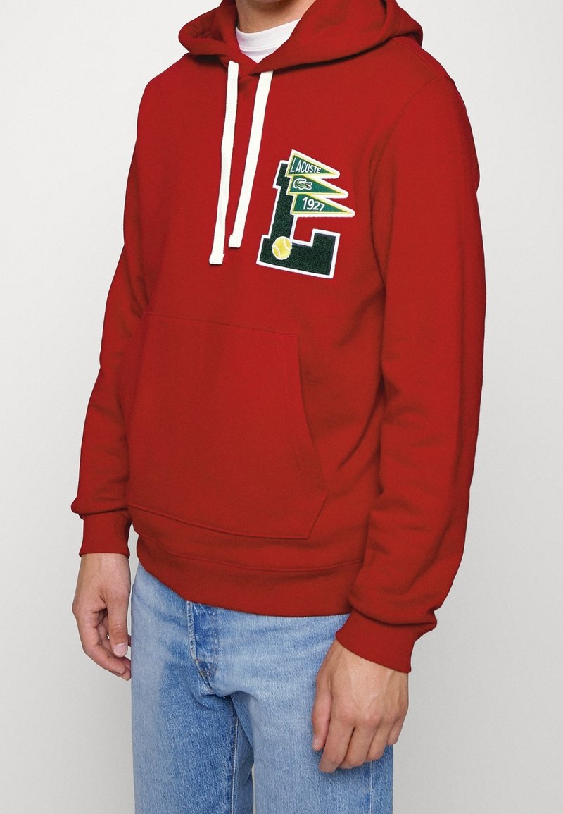 Homme portant un sweat à capuche rouge avec le logo Lacoste et un jean bleu sur un fond uni.