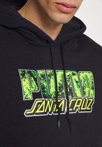 Felpa nera con grafica frontale, caratterizzata da un testo in grassetto "PUMA" in verde con un motivo marmorizzato e "SANTA CRUZ" sottostante in bianco e dettagli gialli.
