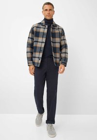 Geruite shirt in blauw, grijs en beige, met knopen en een kraag. Draagt over een donkere coltrui, gecombineerd met marineblauwe broek en grijze sneakers.