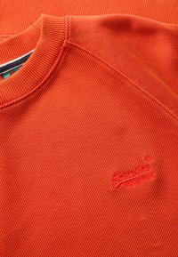 Pomarańczowy sweter z prążkowanej tkaniny, z fakturowaną powierzchnią, okrągłym dekoltem i haftowanym logo na dolnej lewej części.