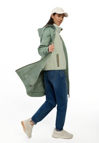 Mintgroene waterdichte jacket over een lichtgroene fleece, gecombineerd met blauwe jeans en beige sportschoenen. Beschikt over een capuchon en ritssluiting.