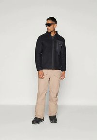 Must have mustrevärviline fliistekk, millel on lukuga esiosa ja rinna tasku; koos hallide cargo pükste ja mustade kingadega. Mudel kannab prille.