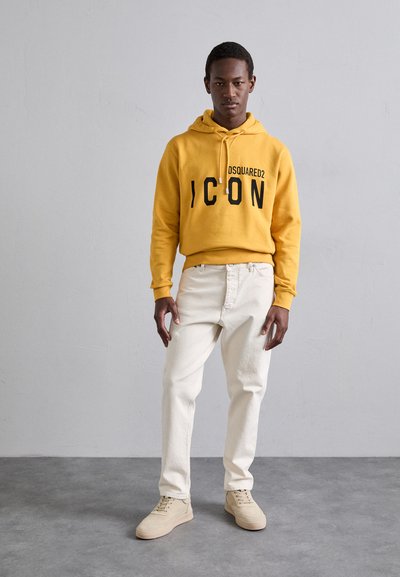 Felpa con cappuccio gialla e testo nero, jeans beige chiaro e sneakers beige. Design semplice, tessuto morbido e stile minimalista.
