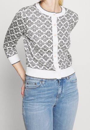 Femme portant un cardigan blanc avec des motifs floraux noirs et des boutons blancs, associé à un jean bleu clair, la main reposant sur sa hanche.