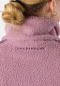 Roze fleece jack met een hoge kraag en een gestructureerde afwerking, met "SWEDEMOUNT" in donkerpaars geborduurd op de achterkant.