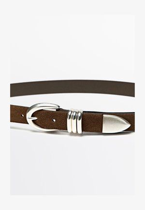 Brązowy pasek zamszowy z metalową, srebrną klamrą, trzema dekoracyjnymi srebrnymi kółkami oraz zakończeniem w kształcie grotu do zapięcia. Widoczna faktura powierzchni.