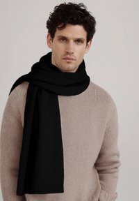 Écharpe noire côtelée à la texture lisse, élégamment drapée autour du cou, associée à un pull beige en tricot.