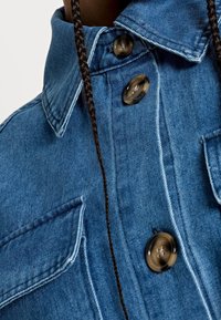 Kurtka dżinsowa w jasnoniebieskim kolorze, z ostro zakończonym kołnierzem, dwoma kieszeniami na piersi oraz dużymi brązowymi guzikami. Prosty krój z teksturowanym wykończeniem.