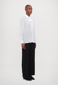 Vit, genomskinlig blus med lång ärm och en slipsdetalj vid kragen, i kombination med vida svarta byxor och eleganta svarta klackstövlar.