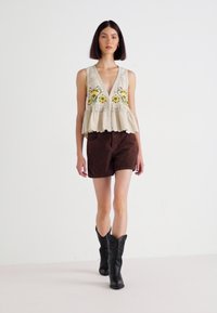 Top sin mangas bordado en color marrón con diseño floral, combinado con pantalones cortos de ante marrón y botas vaqueras negras, sobre un fondo liso.