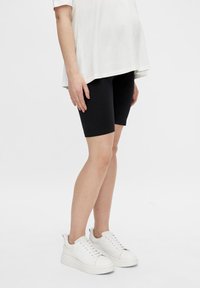 Shorts de cyclisme noirs en tissu extensible, associés à un haut blanc ample et des baskets blanches, sur un fond uni.