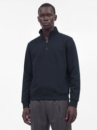 Homme portant un pull noir à fermeture éclair quart, et un pantalon gris, debout devant un fond blanc.