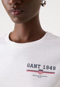 Biała bawełniana koszulka T-shirt z nadrukiem "GANT 1949" w kolorach granatowym, czerwonym i białym. Okrągły dekolt, gładka tekstura i akcent logo.