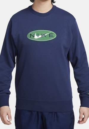 Homme portant un sweat-shirt bleu marine à manches longues avec un logo Nike ovale vert avec un swoosh blanc sur la poitrine, les mains détendues sur les côtés.