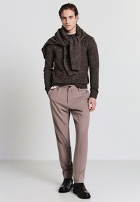 Brauner strukturierter Strickpullover mit einem über die Schultern drapierten Schal, kombiniert mit hellbeigen maßgeschneiderten Hosen und schwarzen Lederstiefeln.