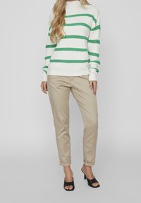 Femme portant un pull blanc à rayures horizontales vertes, un pantalon beige retroussé et des sandales à talons ouvertes noires, debout devant un fond gris.