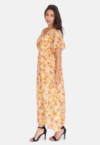 Abito maxi floreale con maniche corte in morbido tessuto pesca, decorato con fiori gialli e arancioni, vitacinchata, gonna fluida, indossato con tacchi neri.