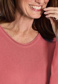 Nahaufnahme einer lächelnden Frau, die einen rosa Pullover trägt und mit ihrer rechten Hand in der Nähe ihres Mundes ihr Gesicht berührt.